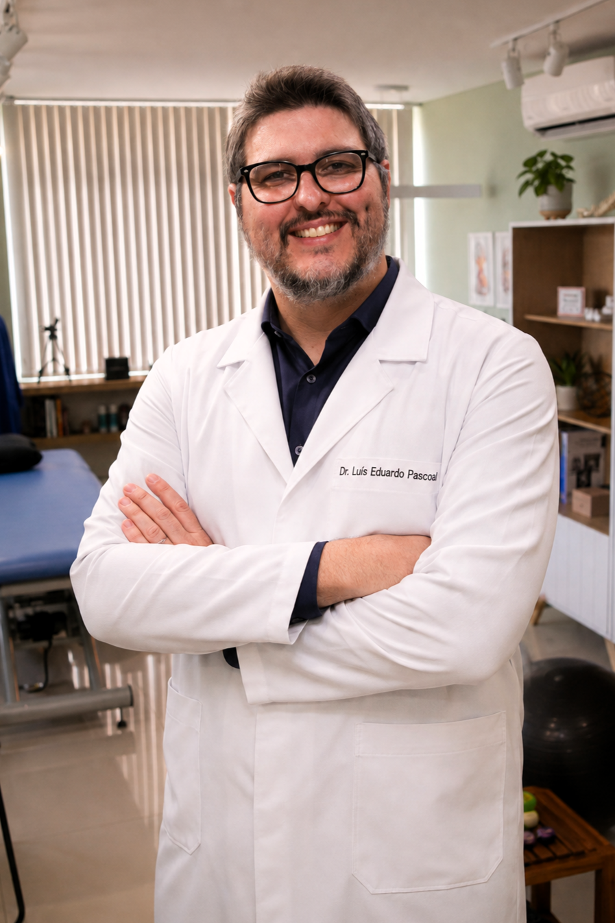 Retrato do Dr. Luís Eduardo Pascoal, fisioterapeuta da EARFF, de jaleco branco com o nome bordado, óculos de grau, braços cruzados, sorrindo, em consultório clínico com maca de fisioterapia azul ao fundo, iluminação natural suave, fotografia corporativa realista