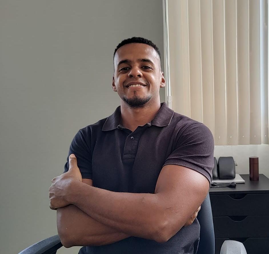 Retrato do Dr. Raphael Nunes, fisioterapeuta da EARFF, sorrindo com braços cruzados, vestindo polo azul-marinho, sentado em cadeira de escritório, ao fundo persiana clara e móvel escuro com telefone e acessórios, iluminação natural suave, fotografia corporativa realista