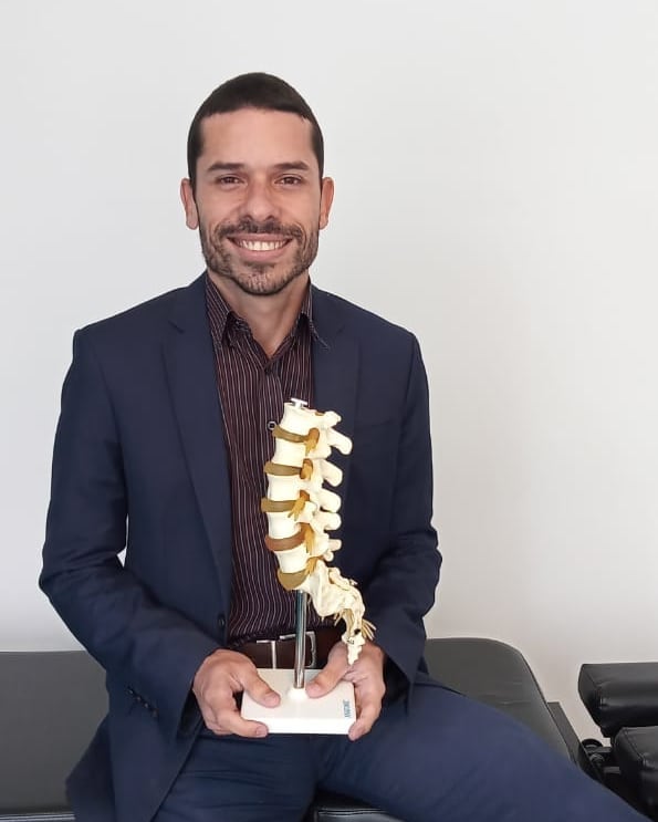 Retrato do Dr. Angel Mello, fisioterapeuta da EARFF, sorrindo, vestindo terno azul-marinho e camisa listrada, segurando modelo anatômico de coluna vertebral, sentado em ambiente clínico com sofá cinza, iluminação natural suave, fotografia corporativa realista
