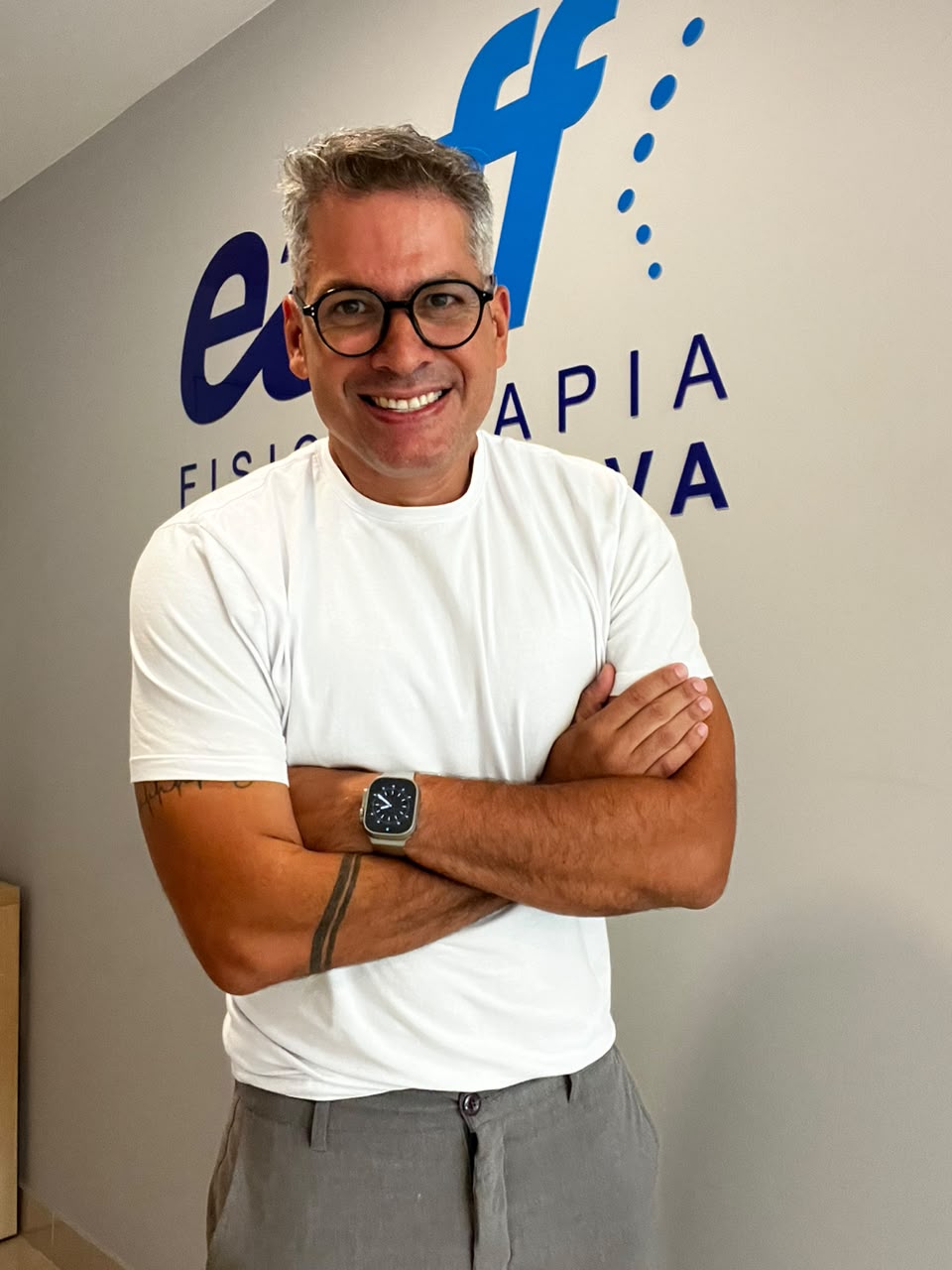 Retrato do Dr. Fernando Capristano, fisioterapeuta da EARFF, sorrindo com braços cruzados, vestindo camiseta branca, tatuagem no antebraço, relógio no pulso, ao fundo parede com logo da clínica EARFF Fisioterapia Investigativa em azul, iluminação natural, fotografia realista