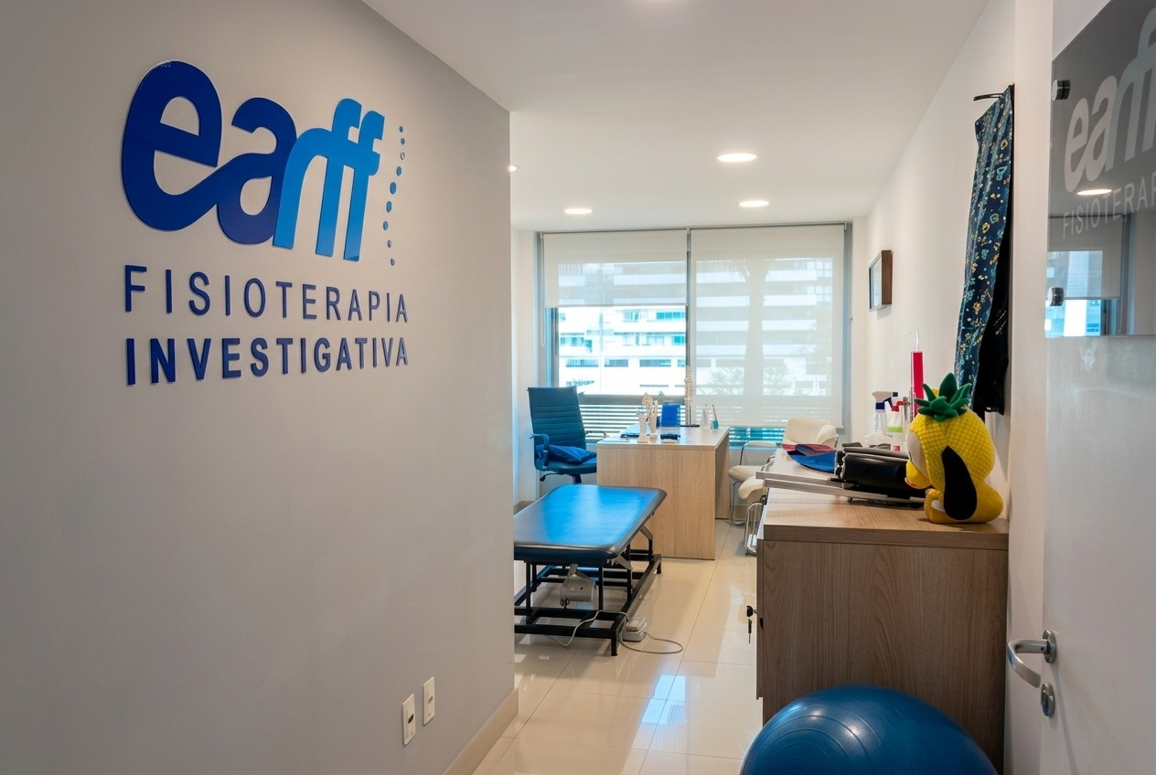 Corredor de acesso à sala de atendimento da unidade EARFF Freguesia, parede com logo EARFF Fisioterapia Investigativa em azul, maca de fisioterapia azul, mesa de madeira com materiais de atendimento e mascote pinguim de pelúcia, janela com persiana e vista para prédios, piso de porcelanato claro, ambiente clínico moderno e iluminado, fotografia profissional realista
