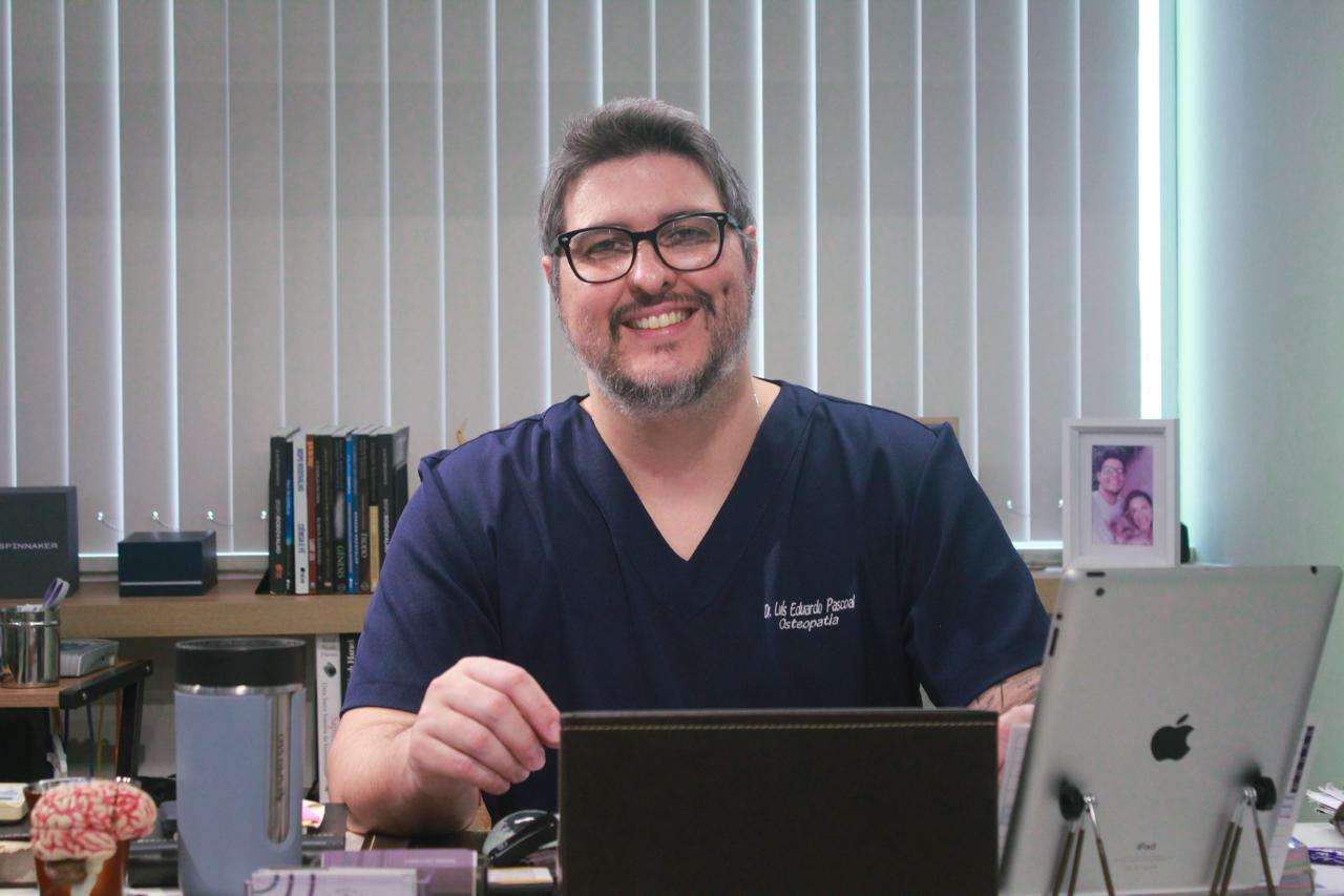 Retrato do Dr. Luís Eduardo Pascoal, fisioterapeuta da EARFF, sorrindo em seu consultório, vestindo scrub azul-marinho bordado com seu nome e especialidade em osteopatia, óculos de grau, ambiente de escritório clínico com persianas verticais ao fundo, iluminação natural suave, fotografia realista
